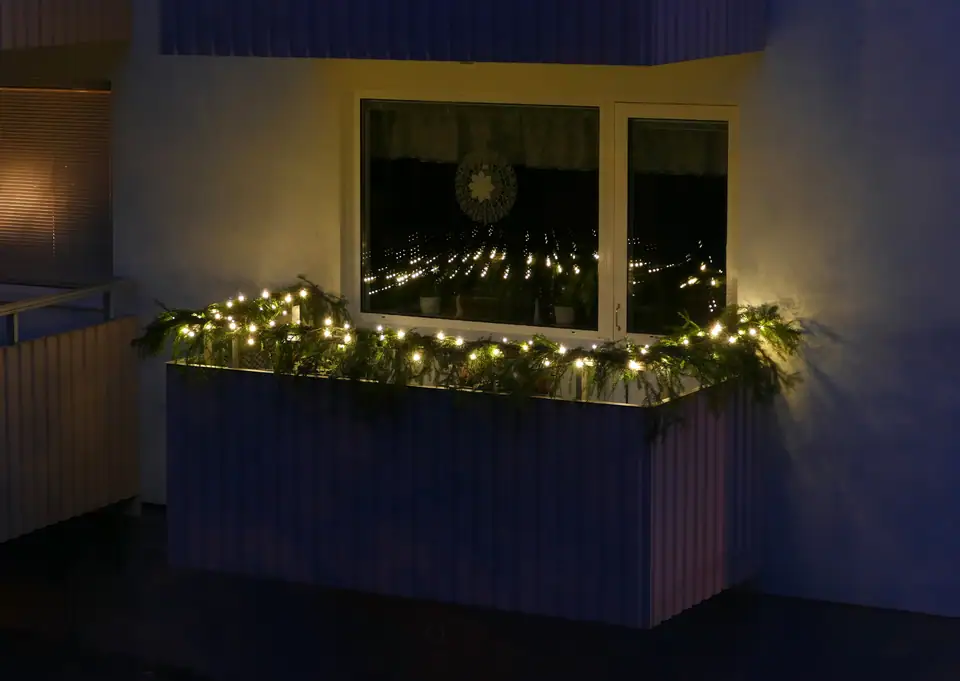 Warm white LED string lights wound through spruce twigs along an apartment balcony railing at night