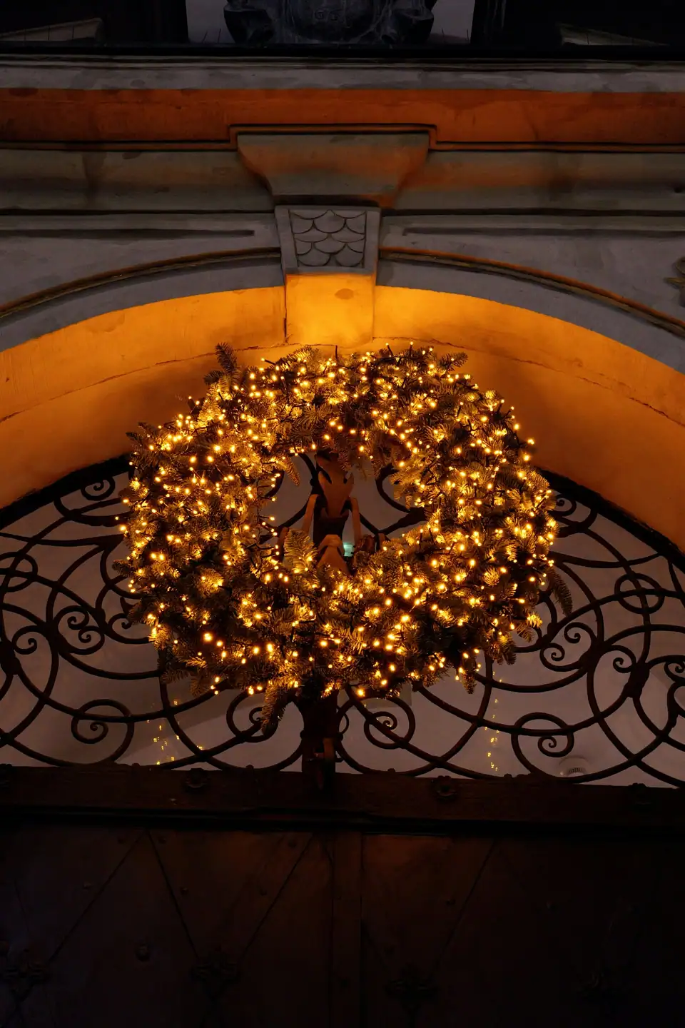 A wreath wrapped in warm white lights hanging in an arched front doorway with an iron grille