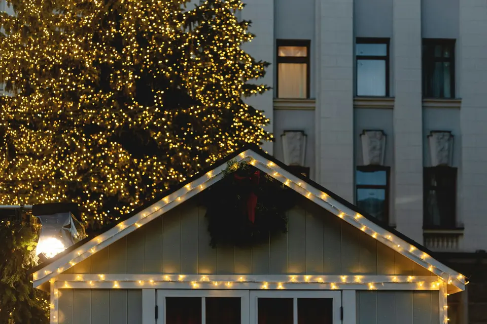 A small building with warm lights along its roofline and a wreath, beside a large tree covered in golden lights