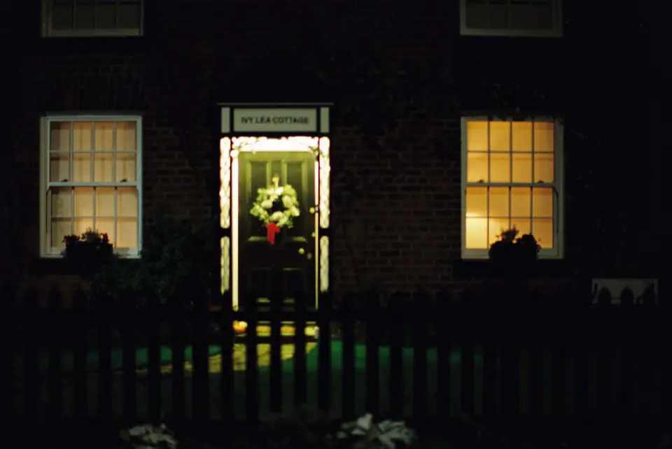 A Christmas wreath on a front door in a lit, covered doorway at night, with glowing windows on either side