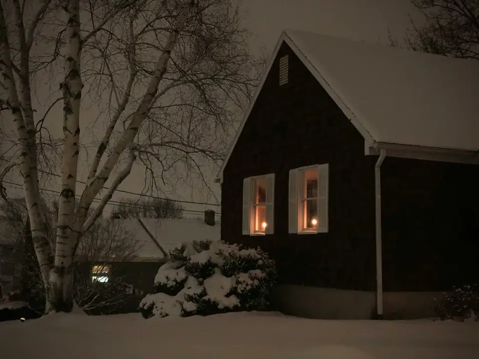 An old house under fresh snow at night, with lit candles glowing in its front windows